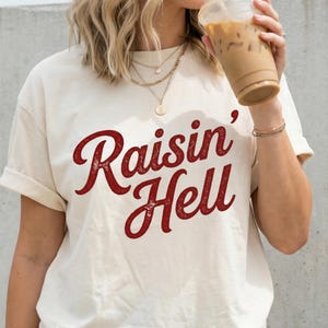 May include: A cream-colored short-sleeve t-shirt featuring the text Raisin' Hell printed in a distressed, dark red script font across the chest. The shirt has a crew neckline and a relaxed fit, worn with layered gold necklaces and a bracelet.