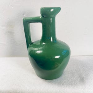 May include: A glossy, emerald green ceramic pitcher with a handle and a spout. The pitcher has a rounded body and a square handle. The spout is slightly pointed. The pitcher is a decorative item.