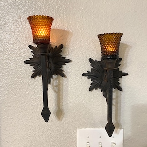 May include: Two wall-mounted sconces featuring dark metal backplates with a leaf-like design and long, pointed stems. Each sconce holds a glowing amber-colored glass shade with a textured hobnail pattern, emitting a warm light.