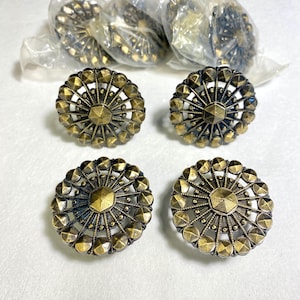 May include: Antique brass-colored cabinet knobs with a starburst design. The knobs have a central faceted shape surrounded by a detailed pattern of small, raised geometric shapes. Several knobs are visible, some still in their original packaging.