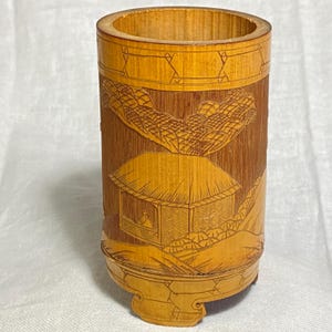 May include: A cylindrical bamboo brush pot featuring intricate carved details of a small hut with a thatched roof, surrounded by stylized landscape elements. The natural light brown wood grain is visible throughout the surface of the vessel.