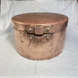 May include: A round copper container with a lid, featuring a weathered, oxidized surface with dark spots and a patina. The lid is secured by a central metal hinge mechanism with four visible rivets, showing signs of age and use throughout the metal.
