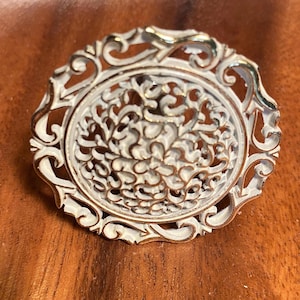 May include: An oval-shaped brooch with a white filigree design and gold-colored trim. The brooch features an intricate, openwork pattern in the center, surrounded by a decorative border. The overall design is ornate and vintage-inspired.