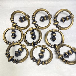 May include: A collection of nine antique brass drawer pulls. Each pull features a circular handle with a decorative center piece and a square mounting plate. The pulls have a vintage, aged appearance.