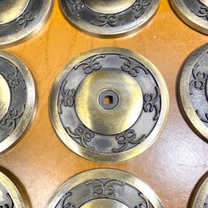 May include: Close-up of several antique brass-colored metal coins. Each coin has a central square hole and is decorated with an ornate, symmetrical design around the edges. The coins are arranged on a light brown surface.