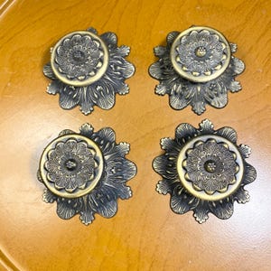 May include: Four antique brass cabinet knobs with a floral design. Each knob features a central circular element with a detailed flower pattern, surrounded by a stylized, petal-like base. The knobs have a vintage aesthetic.