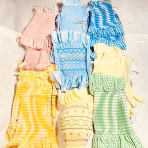 May include: A collection of rectangular fabric panels featuring smocking and ruffles in various colors including pink, light blue, yellow, and green. Some panels display gingham patterns, while others show intricate decorative stitching in contrasting thread.