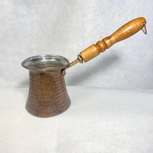 May include: A hammered copper coffee pot with a wide, flared rim and a pouring spout. The pot features a long, turned wooden handle attached with a brass-colored metal fitting. A small metal loop is attached to the end of the wooden handle.