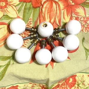 May include: A collection of white ceramic cabinet knobs with brass and silver hardware. The knobs are round and smooth, and the hardware includes threaded rods and nuts. The knobs are arranged on a floral patterned fabric.