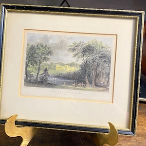 May include: A framed landscape print titled UGBROOKE, DEVONSHIRE, featuring a yellow manor house, a lake, trees, and deer. The artwork is enclosed in a black and gold frame with a cream-colored mat, presenting a detailed historical scene.