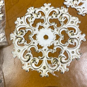 May include: White and gold filigree decorative hardware. The ornate, circular design features a central hole and intricate scrollwork. The hardware has a distressed finish, with gold accents on the raised details. The hardware is likely used for furniture or decorative purposes.