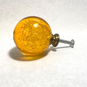 May include: A decorative cabinet knob with a clear, amber-colored glass sphere filled with gold glitter. The knob has a brass base and a silver screw for attachment. The overall design is a round shape.