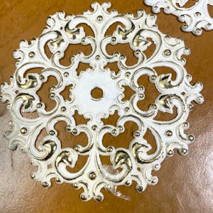 May include: Ornate, white, circular decorative elements with gold accents. The design features swirling patterns and small, raised gold details. Each piece has a central hole and is set against a brown background.