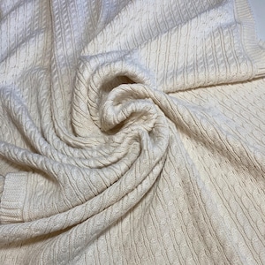 May include: Cream-colored cable knit blanket. The blanket is folded and draped, showcasing the intricate woven pattern. The soft, textured material is visible throughout the image, with a focus on the detailed cable design. The blanket appears cozy and warm.