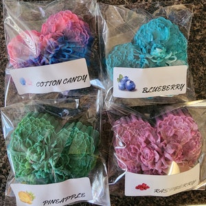 Puede incluir: Cuatro jabones en forma de corazón con texturas florales intrincadas, envueltos individualmente en plástico transparente. Los jabones son rosa y azul, azul brillante, verde y morado. Cada etiqueta dice Cotton Candy, Blueberry, Pineapple o Raspberry.