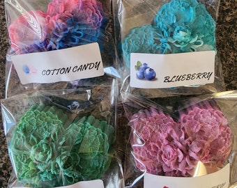 May include: Four heart-shaped, textured floral soaps individually wrapped in clear plastic. The soaps are coloured pink and blue, blue, green, and purple. Each has a white label with text: Cotton Candy, Blueberry, Pineapple, and Raspberry.