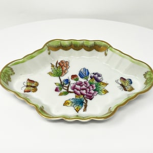May include: A small, white porcelain dish with a green and gold rim. The dish features a floral design with butterflies in shades of pink, purple, yellow, and green.