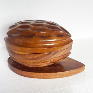 Peut inclure: Une boîte en bois en forme de noix avec un couvercle texturé et une base séparée. La boîte est en bois brun foncé et a une finition naturelle.