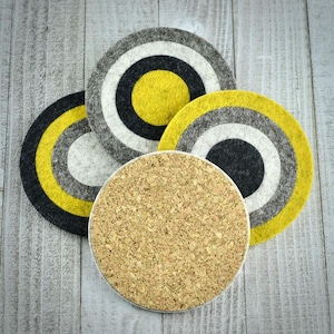 May include: Four round coasters. Three are made of felt with concentric circles in gray, white, black, and yellow. One coaster is made of cork. The coasters are arranged on a light gray wooden surface.