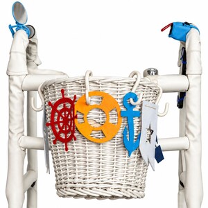 May include: A white wicker basket attached to a walker frame, decorated with felt nautical shapes including a red ship wheel, an orange life preserver, a blue anchor, and a white pennant with a star cutout, plus a small round mirror.
