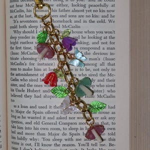 May include: A decorative keychain with a gold-colored clasp and chain. The chain is adorned with colorful beads, including flowers, butterflies, and leaves. The keychain is set against the backdrop of an open book.