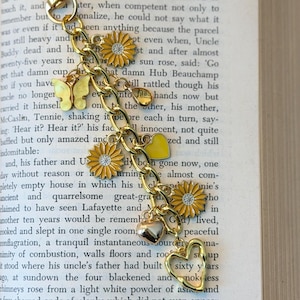 May include: Gold-toned charm bracelet with daisy, butterfly, and heart charms. The bracelet is attached to a gold-colored clasp. The charms are yellow, gold, and silver. The bracelet is resting on the pages of an open book.