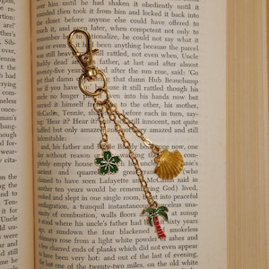 May include: Gold-toned keychain with a lobster clasp, featuring charms of a green flower, a seashell, and a palm tree with a red stripe. The keychain is attached to a book.