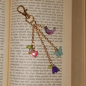May include: Gold-toned keychain with a lobster clasp, adorned with charms including a pink heart, a purple flower, a butterfly, and a crescent moon. The charms are connected by delicate chains.