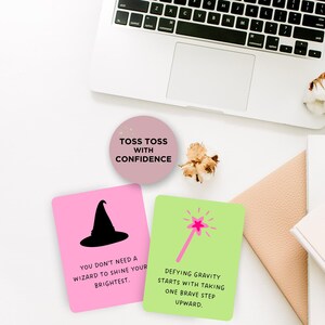 May include: Two pink and green affirmation cards with inspirational quotes about confidence and defying gravity. The cards feature a black witch hat and a pink magic wand with a star on top. The cards are on a white surface with a laptop, a notebook, and a cotton plant.