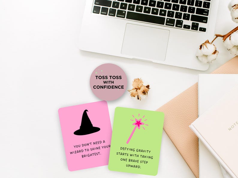 May include: Two pink and green affirmation cards with inspirational quotes about confidence and defying gravity. The cards feature a black witch hat and a pink magic wand with a star on top. The cards are on a white surface with a laptop, a notebook, and a cotton plant.
