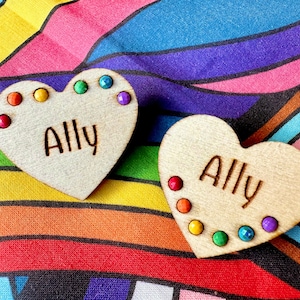 Pode incluir: Dois ornamentos de madeira em forma de coração com a palavra "Ally" gravada neles. Cada coração é decorado com gemas de cores do arco-íris.