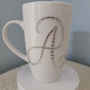 Può includere: Tazza in ceramica bianca con un design a forma di lettera "A" in strass argentati sul davanti.