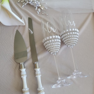 May include: A cake knife and server set with white handles embellished with pearls and rhinestones, paired with two clear champagne flutes featuring white script text reading Mr and Mrs, decorated with rows of pearls and rhinestones at the base.