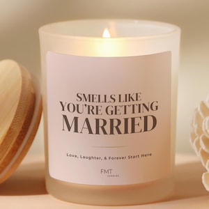 May include: A lit candle in a frosted glass jar with a label that reads "SMELLS LIKE YOU'RE GETTING MARRIED." The text "Love, Laughter, & Forever Start Here" is below the main text. The candle is a wedding-themed product.