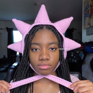May include: A pink crocheted hat with a star-shaped design. The hat has a long, pointed crown and a wide brim. The hat is worn by a person with black braids.