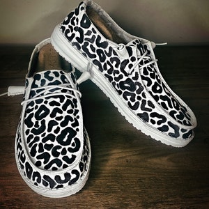 May include: A pair of white canvas slip-on shoes with a black leopard print design.