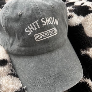 May include: Gray baseball cap with the words "SHIT SHOW SUPERVISOR" printed in white. The hat has a curved brim and a fabric strap closure. The hat is made of a soft, worn material.