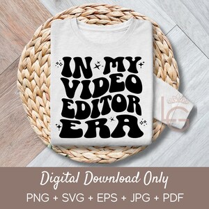 Op de afbeelding: Een wit T-shirt met zwarte tekst die "IN MY VIDEO EDITOR ERA" luidt in een retro-lettertype. De tekst is omringd door kleine sterren. Het T-shirt is gevouwen en geplaatst op een geweven rieten oppervlak.