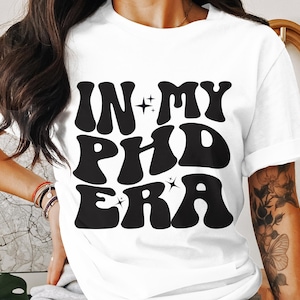 May include: White t-shirt with black text that reads "In My PhD Era" in a retro font style.