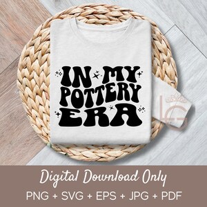 Puede incluir: Una camiseta blanca con el texto "IN MY POTTERY ERA" en letras negras de estilo retro. El texto está rodeado de pequeñas estrellas. La camiseta se muestra sobre una superficie de mimbre tejido.