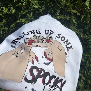 May include: White sweatshirt with a graphic of hands rolling up a collection of horror movie characters. The text reads "Rolling Up Some Spooky Vibes" with bats and stars. The hands have red nail polish.
