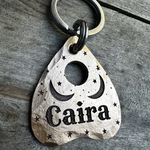 以下が含まれることがあります： 「Caira」の黒い刻印が入った、銀色のハート型のキーホルダー。三日月と星のデザインで、上部に丸い穴があります。シルバーのキーリング付き。