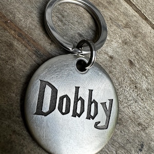 May include: A round, brushed metal keychain tag with the name "Dobby" engraved in a gothic font. The tag is attached to a silver split ring. The background is a wooden surface.