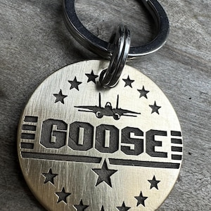 May include: A round, brushed metal keychain with the name "GOOSE" in bold, engraved letters. The keychain features a fighter jet silhouette above the name and is encircled by stars. A silver key ring is attached at the top.