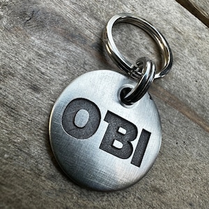 May include: Silver metal dog tag with a black engraved name "OBI" attached to a silver key ring.