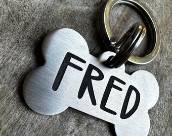 May include: A brushed silver dog tag in the shape of a bone, with the name "FRED" in bold black letters. The tag is attached to a silver split ring. The background is a textured, gray wooden surface.