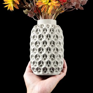 May include: A gray vase with a geometric pattern, filled with colorful artificial flowers. The vase is cylindrical and has a textured surface. The flowers include yellow, orange, and burgundy hues, creating a vibrant contrast against the vase.