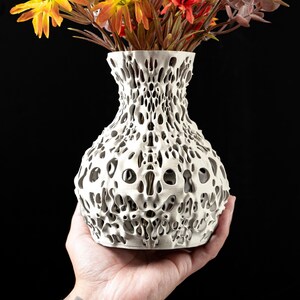 May include: A white, porous vase with a unique, organic design, held in a hand. The vase is filled with colorful artificial flowers in shades of orange, yellow, and red. The vase has a textured, open structure.