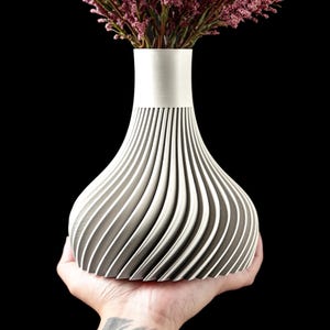 May include: A modern, decorative vase with a unique, ribbed design. The vase is held in a hand and is filled with pink flowers. The vase has a silver top and a gray and white body. The background is black.