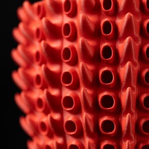 May include: Close-up of a bright red, textured object with a repeating pattern of conical spikes and cylindrical holes. The object has a rough, porous surface and is set against a black background. The design is geometric and visually striking.